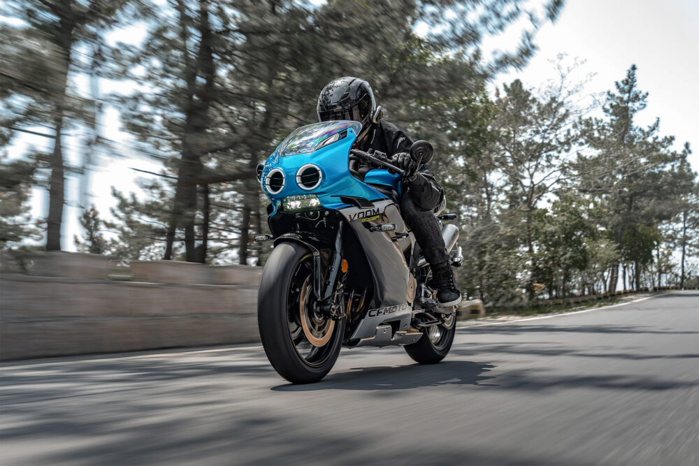 A rider in black gear cruises a blue and silver CFMOTO 500SR VOOM sport motorcycle along a tree-lined road during the day.