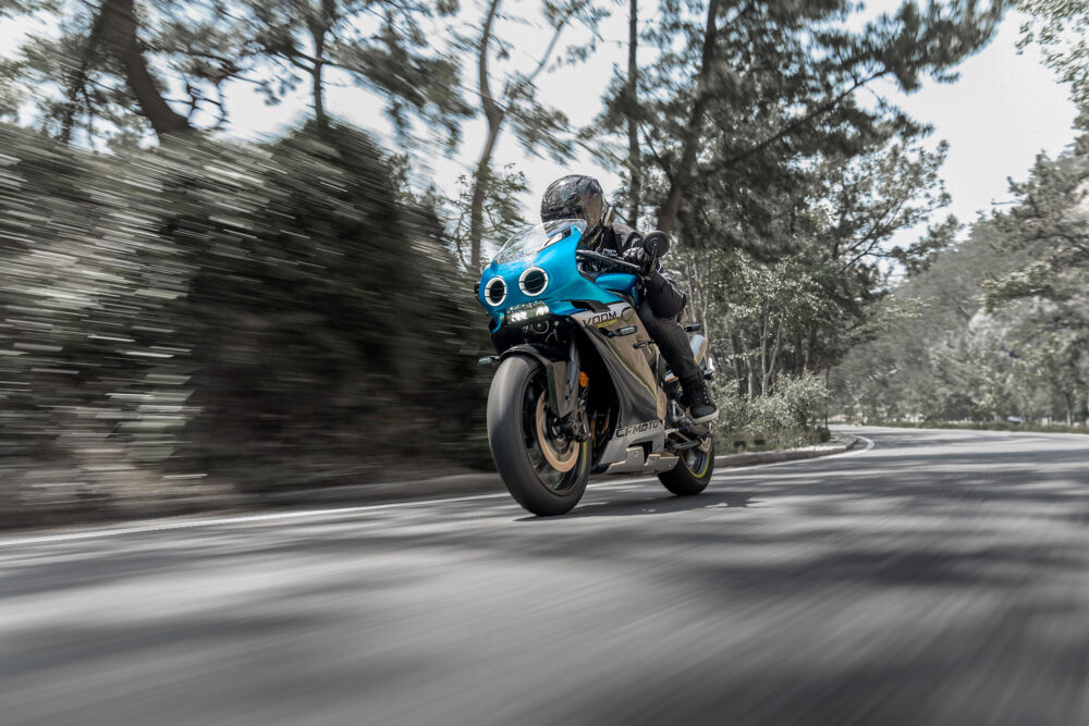 A motorcyclist in black gear rides the CFMOTO 500SR VOOM, a blue sport bike, at speed on a curved road lined with trees during the day.