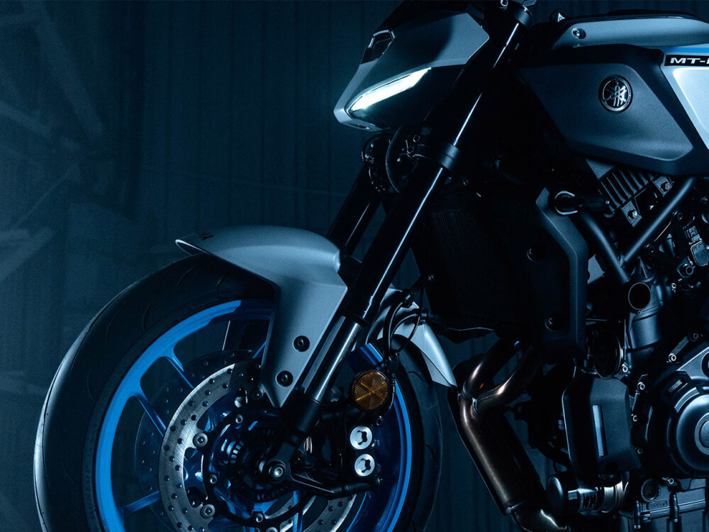 Close-up of the YAMAHA MT-07 HO Y-AMT motorcycle's front wheel and body, featuring blue rims and intricate mechanical details under dramatic lighting.