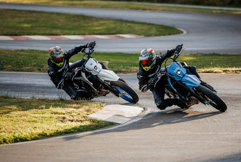 Two motorcyclists in helmets and black gear lean into a racetrack curve, riding SUZUKI DR-Z 4SM motorcycles—one white and one blue.