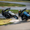 Two motorcyclists in helmets and black gear lean into a racetrack curve, riding SUZUKI DR-Z 4SM motorcycles—one white and one blue.