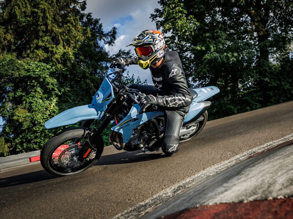 A motorcyclist in full gear leans sharply into a curve on a blue SUZUKI DR-Z 4SM, riding on a paved track bordered by trees and a guardrail.
