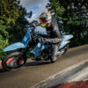 A motorcyclist in full gear leans sharply into a curve on a blue SUZUKI DR-Z 4SM, riding on a paved track bordered by trees and a guardrail.