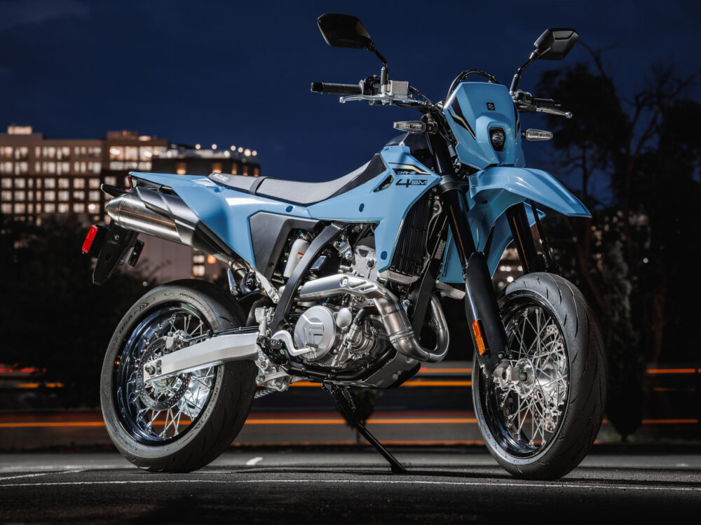A SUZUKI DR-Z 4SM supermoto motorcycle in blue is parked outdoors at night, city buildings glowing in the background.