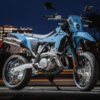 A SUZUKI DR-Z 4SM supermoto motorcycle in blue is parked outdoors at night, city buildings glowing in the background.