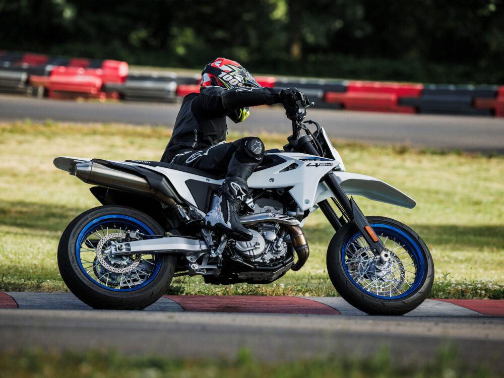 Wearing black riding gear and a helmet, a person leans into a turn on a white SUZUKI DR-Z 4SM motorcycle while riding on a racetrack.