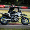 Wearing black riding gear and a helmet, a person leans into a turn on a white SUZUKI DR-Z 4SM motorcycle while riding on a racetrack.