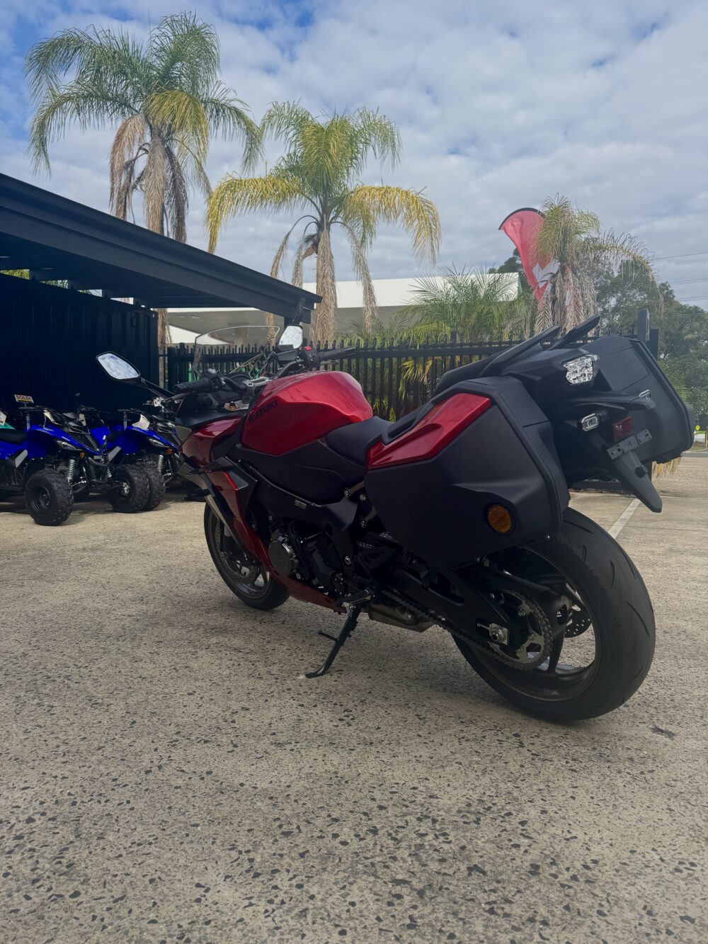 A SUZUKI PANORAMA GT - DEMO sport motorcycle in red and black is parked on a concrete lot near a black fence, with palm trees and ATVs visible in the background.