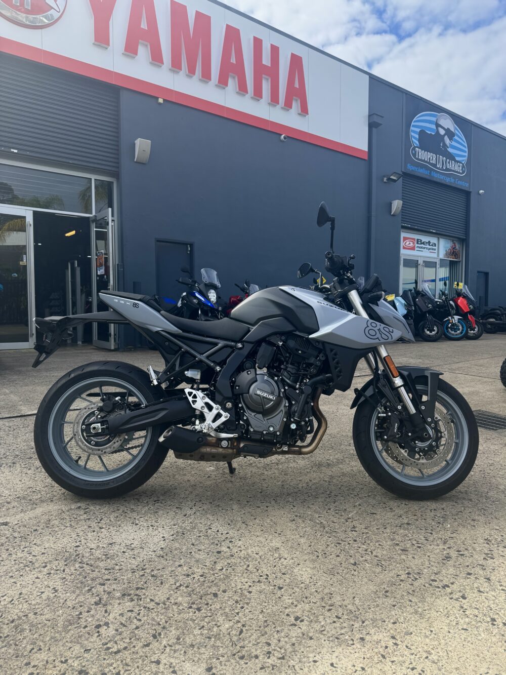 A silver Yamaha motorcycle is parked in front of a Yamaha dealership on concrete, with other bikes—including the SUZUKI GSX-8S DEMO—visible in the background.