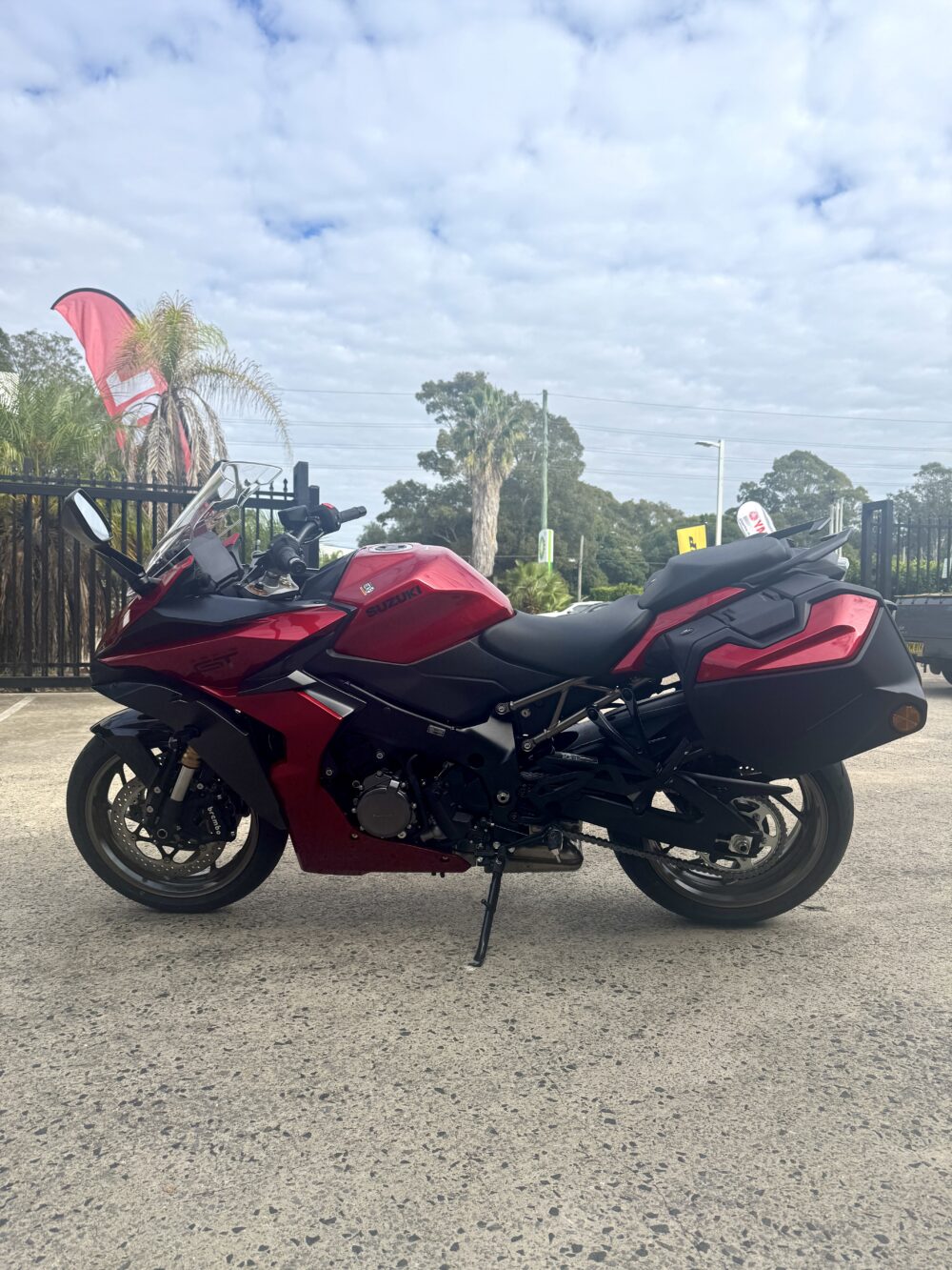 A SUZUKI PANORAMA GT - DEMO in red is parked outdoors on concrete, framed by trees, plants, and a black fence beneath a cloudy sky.