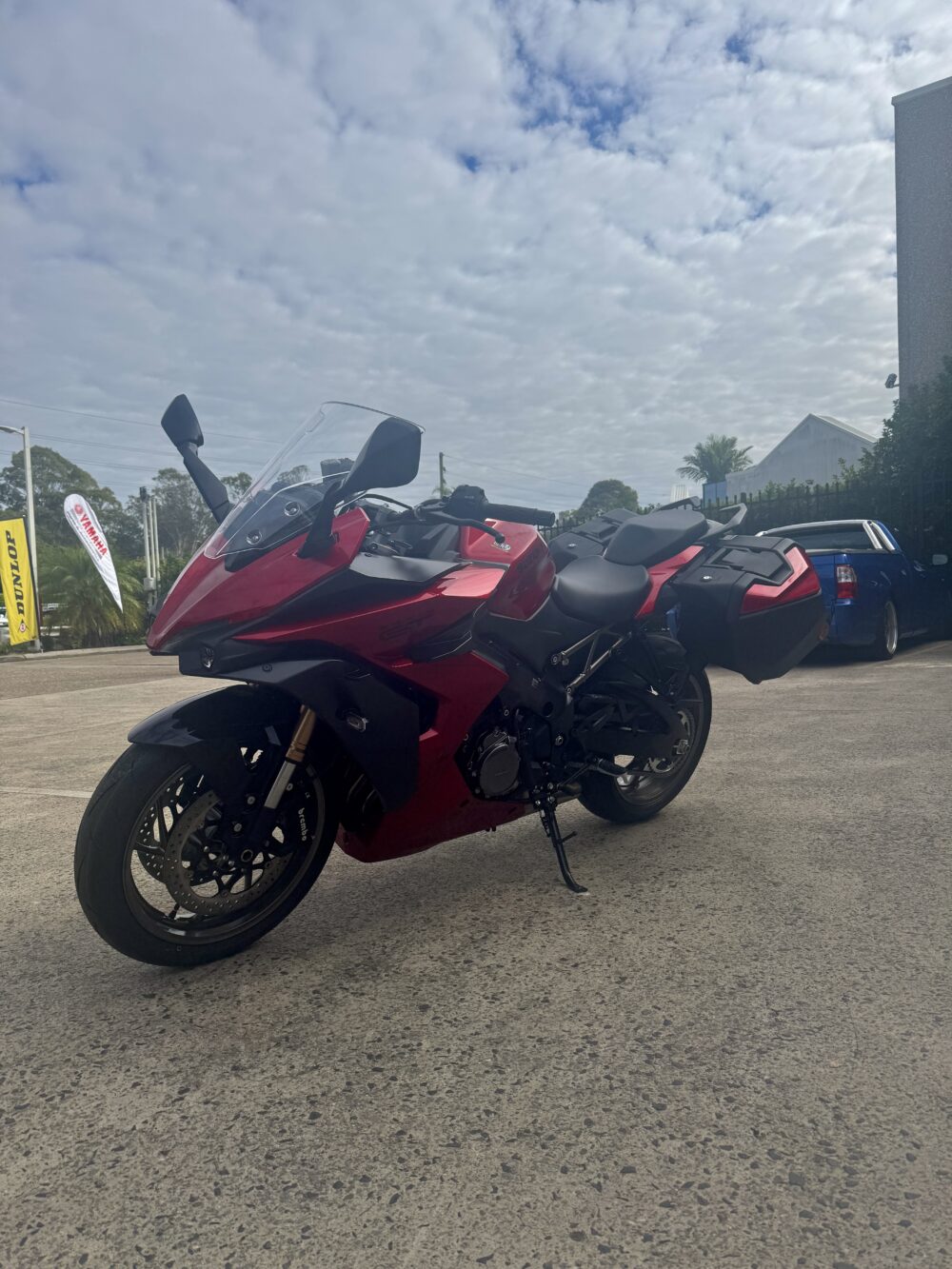 A SUZUKI PANORAMA GT - DEMO sport touring motorcycle in red with saddlebags is parked on concrete under a cloudy sky, near other vehicles.
