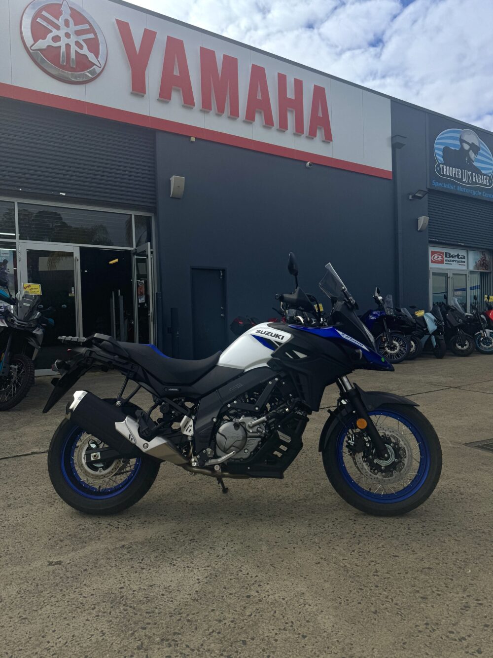 A SUZUKI V-STROM 650XT - DEMO (LAMS) in blue and white is parked in front of a Yamaha dealership building on a cloudy day.