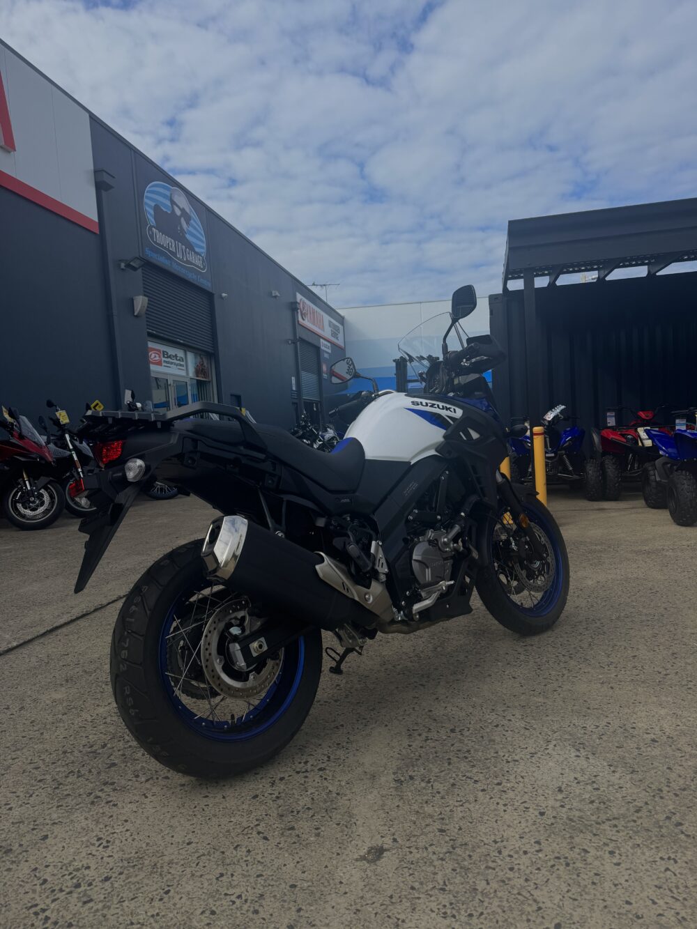 A SUZUKI V-STROM 650XT - DEMO (LAMS) motorcycle in blue and white is parked outside a dealership on a concrete lot beneath a partly cloudy sky.