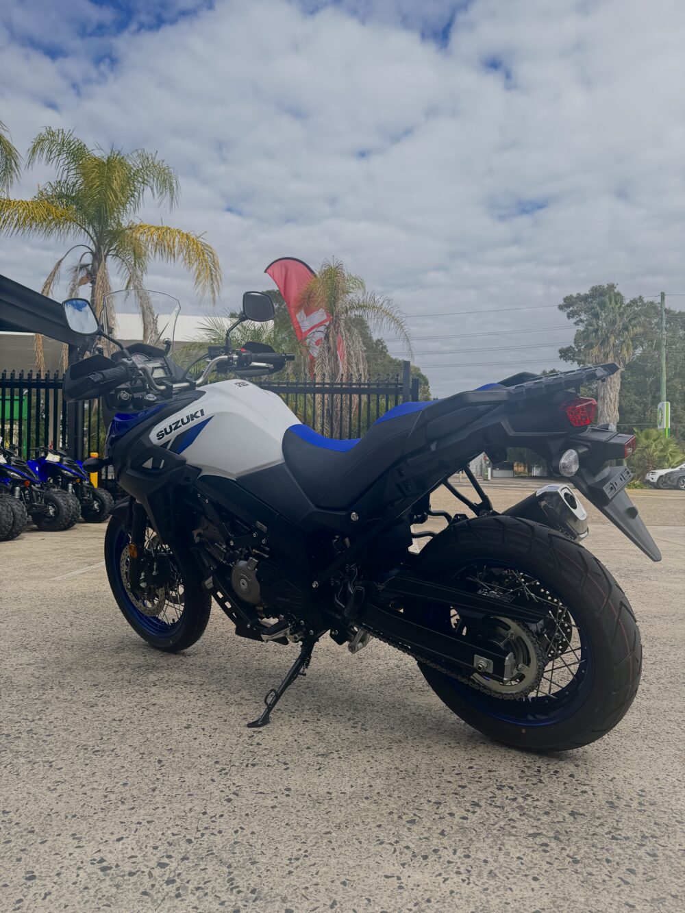 A SUZUKI V-STROM 650XT - DEMO (LAMS) in blue and white is parked on a concrete lot with palm trees and a fence in the background under a partly cloudy sky.