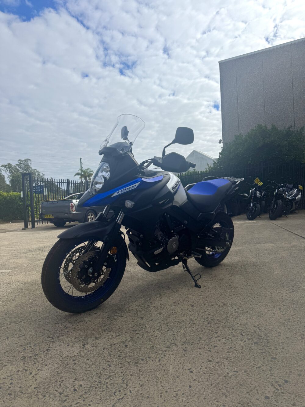 A SUZUKI V-STROM 650XT - DEMO (LAMS) motorcycle in blue and white is parked on a concrete lot near a building, with other motorcycles and greenery visible in the background.