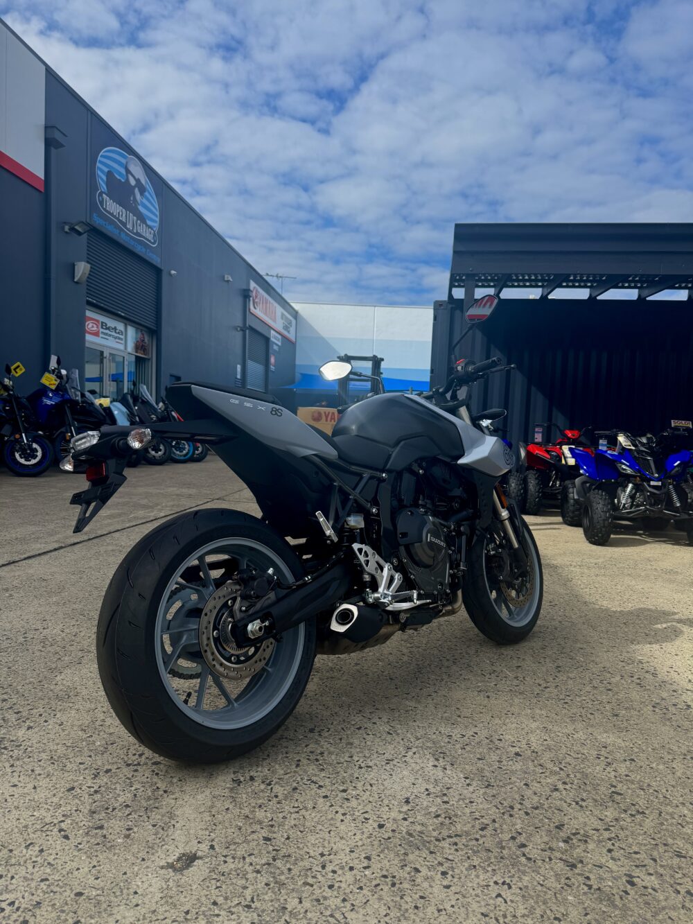 A SUZUKI GSX-8S DEMO in black and white is parked on a concrete lot outside a dealership, with other motorcycles and ATVs in the background beneath a partly cloudy sky.