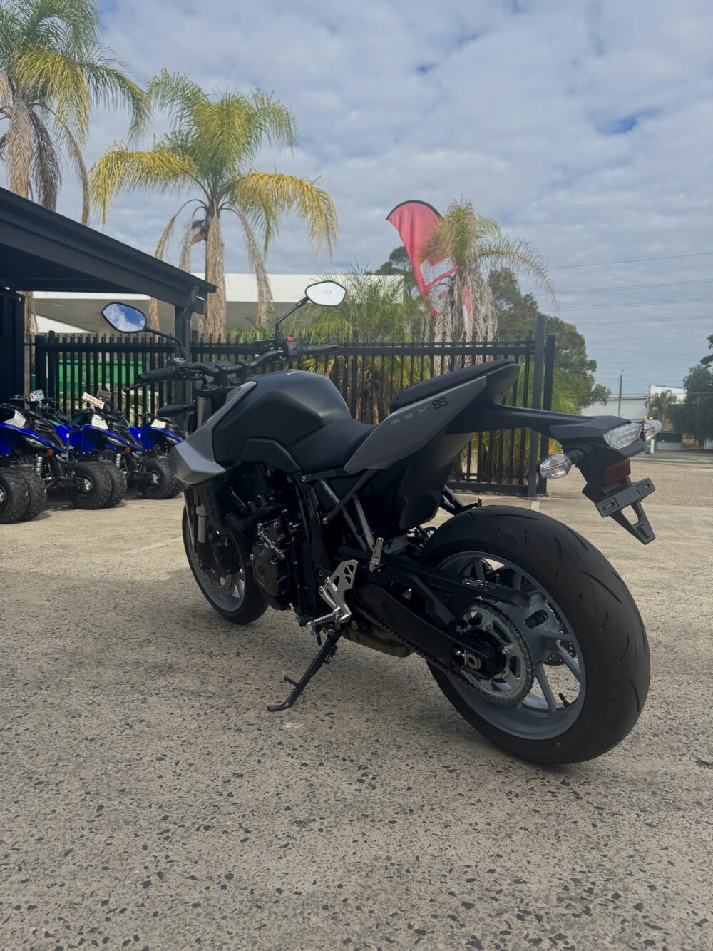 A matte black SUZUKI GSX-8S DEMO is parked on a concrete lot near a fence, with palm trees and blue ATVs visible in the background.