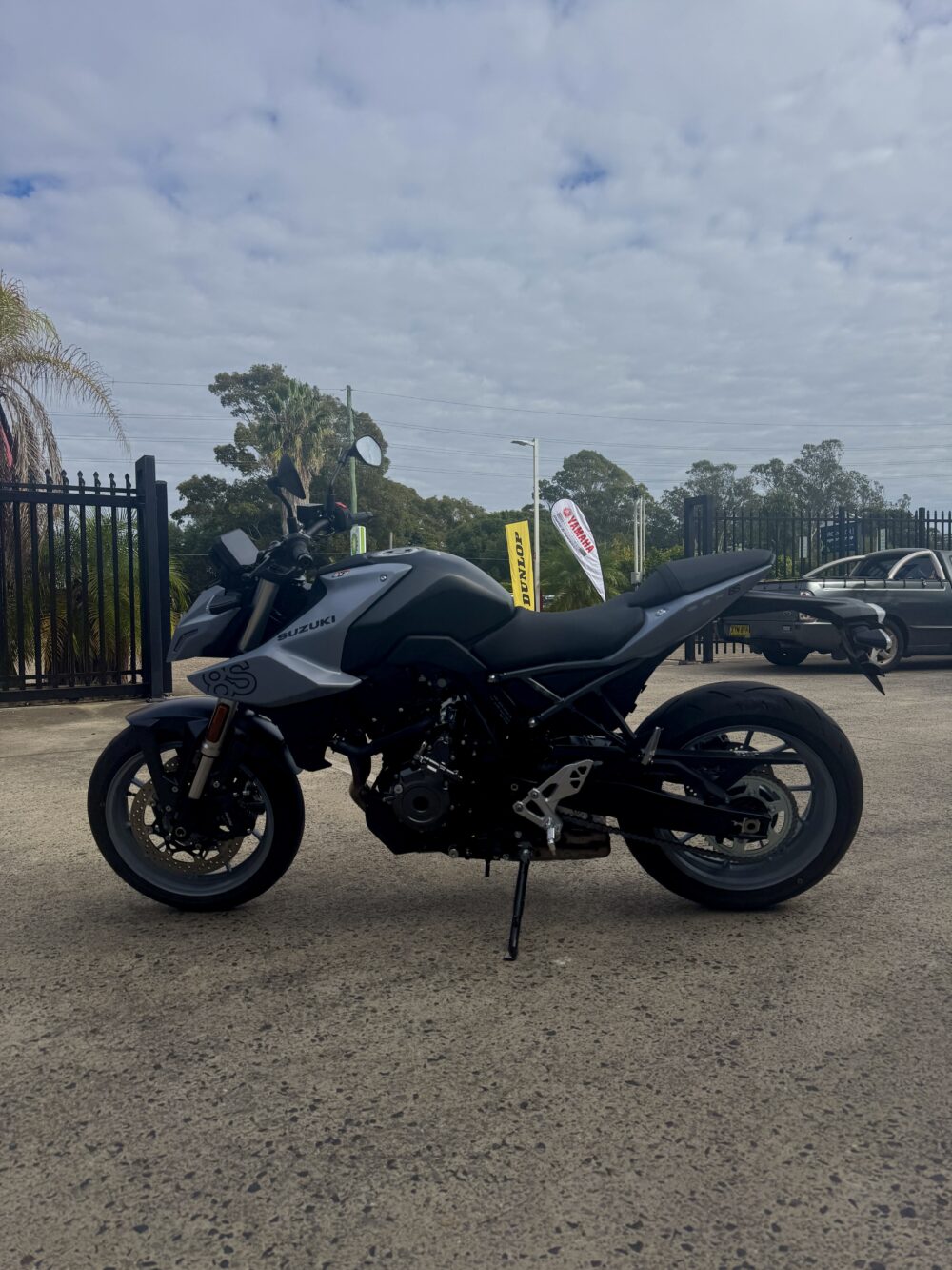 A SUZUKI GSX-8S DEMO motorcycle in black and gray is parked outdoors on pavement, with trees, a black fence, vehicles, and a cloudy sky in the background.