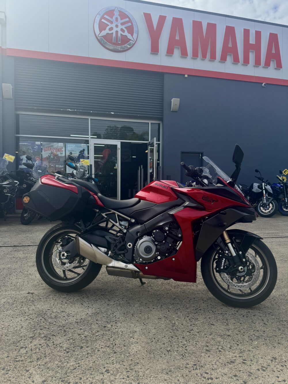 A red Yamaha motorcycle is parked outside a Yamaha dealership, with the store sign above the entrance and other bikes, including a SUZUKI PANORAMA GT - DEMO, visible in the background.
