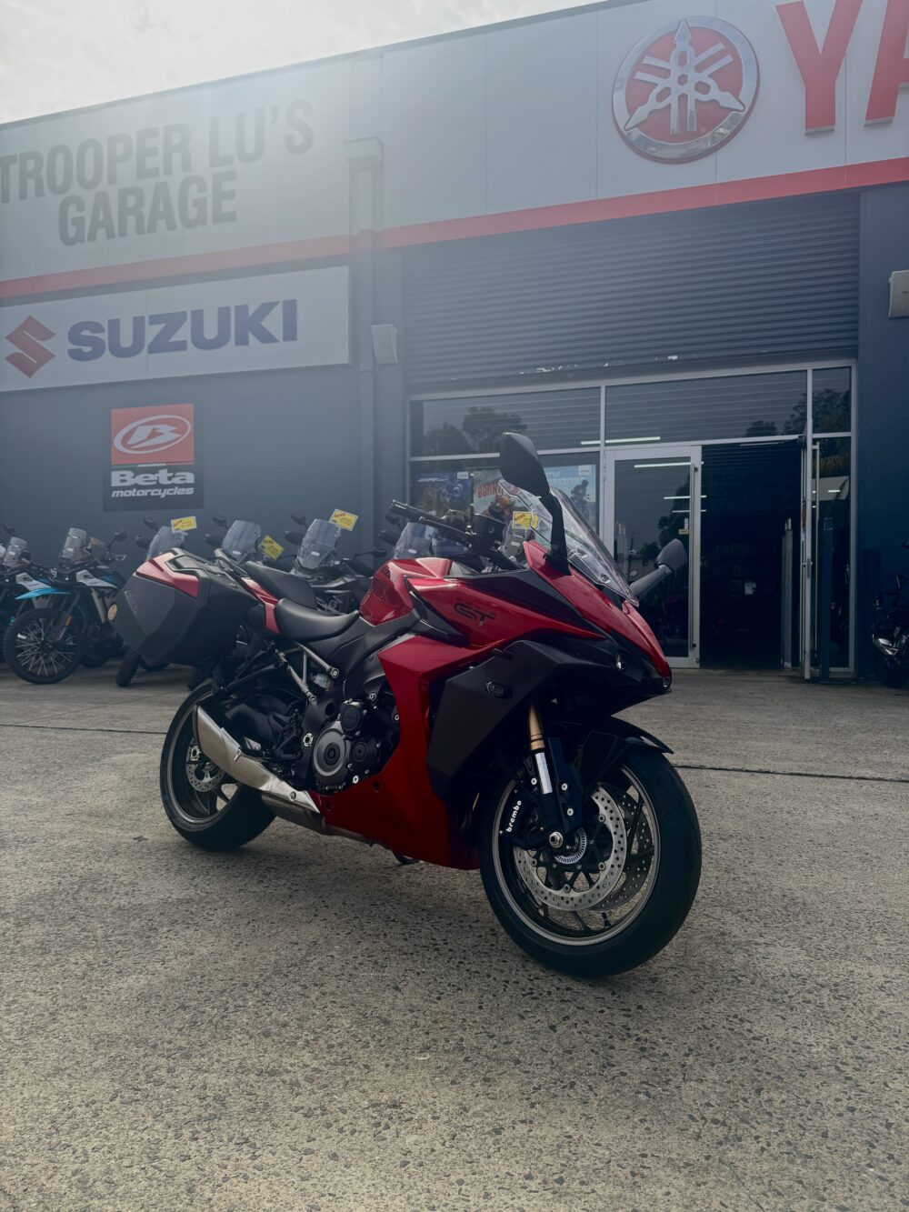 A SUZUKI PANORAMA GT - DEMO sport touring motorcycle in red is parked outside a dealership featuring Suzuki, Beta, and Yamaha branding.